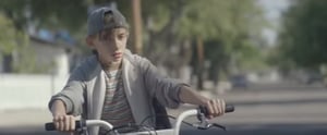 A boy riding home from school encounters a mom who is looking at a post of her beautiful daughter while driving; disaster can be averted if she waits!